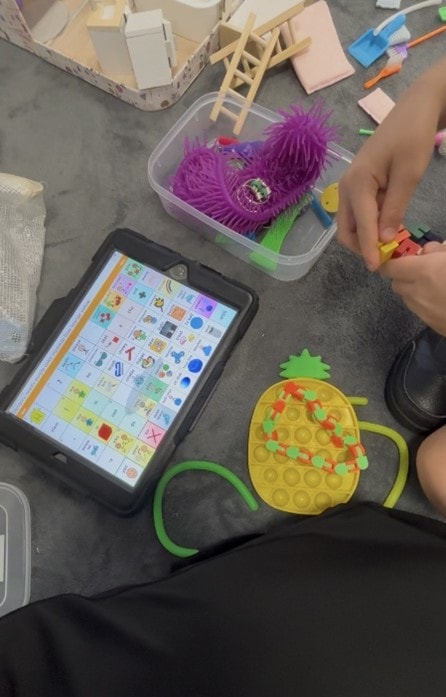 Child using an AAC tablet device with sensory and fine motor play materials during a therapy session